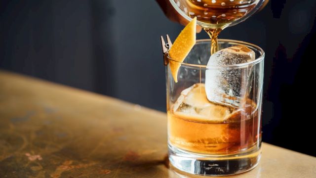 A glass on a bar with ice and an amber drink, garnished with a lemon twist as another drink is poured over it.