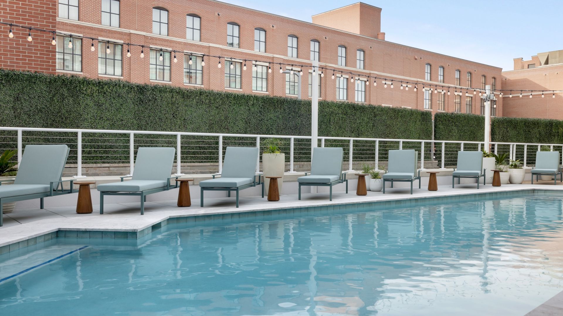 A hotel courtyard pool with lounge chairs, planters, and a brick building with string lights above the hedge-lined patio.