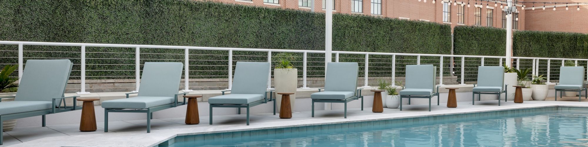 A hotel courtyard pool with lounge chairs, planters, and a brick building with string lights above the hedge-lined patio.