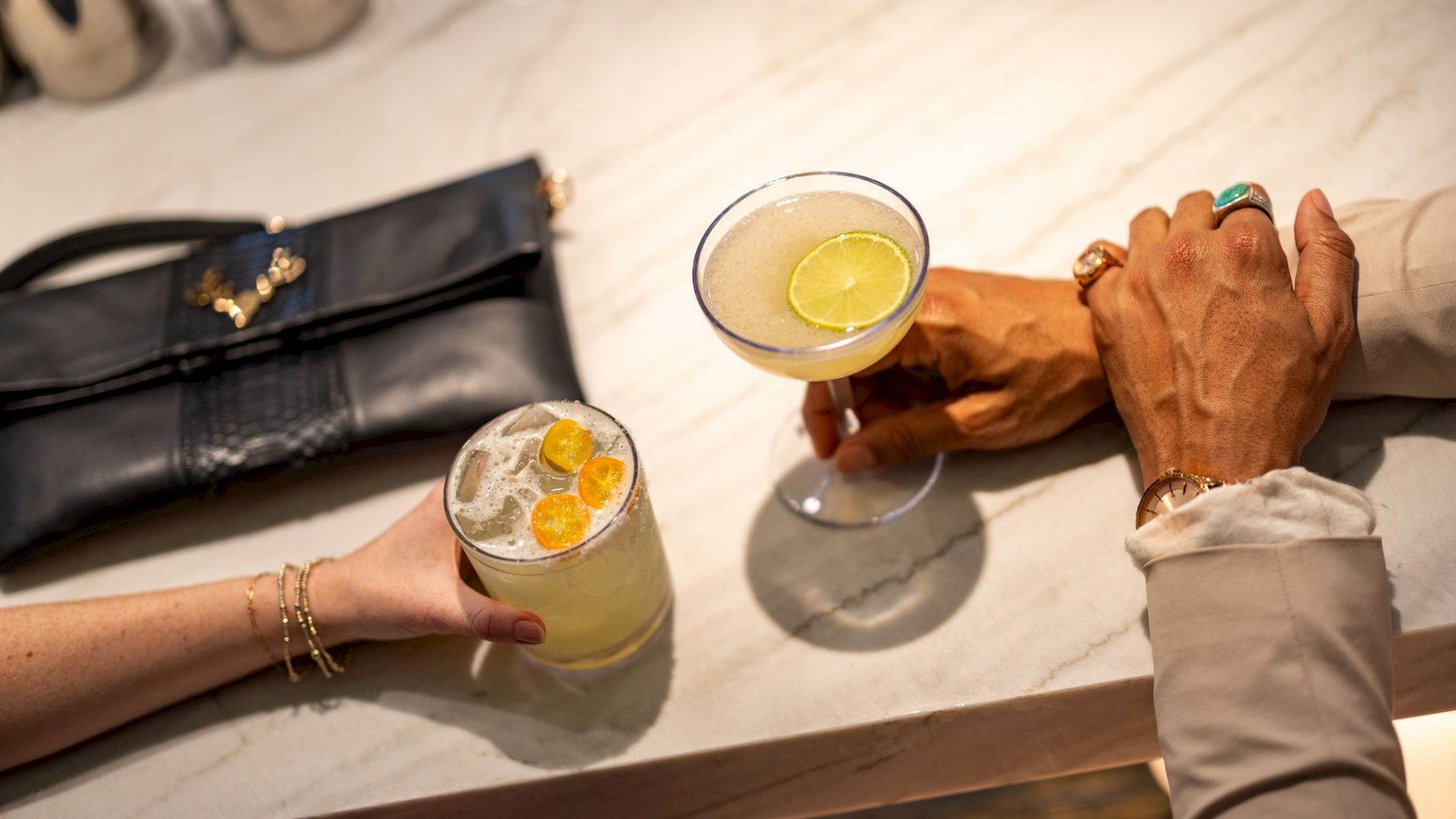 A couple of cocktails on a marble bar, with hands reaching in; a black purse sits nearby. The scene feels like a chic lounge.