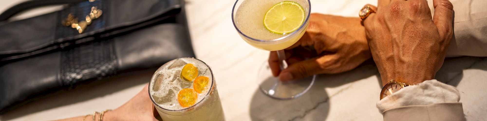 A couple of cocktails on a marble bar, with hands reaching in; a black purse sits nearby. The scene feels like a chic lounge.