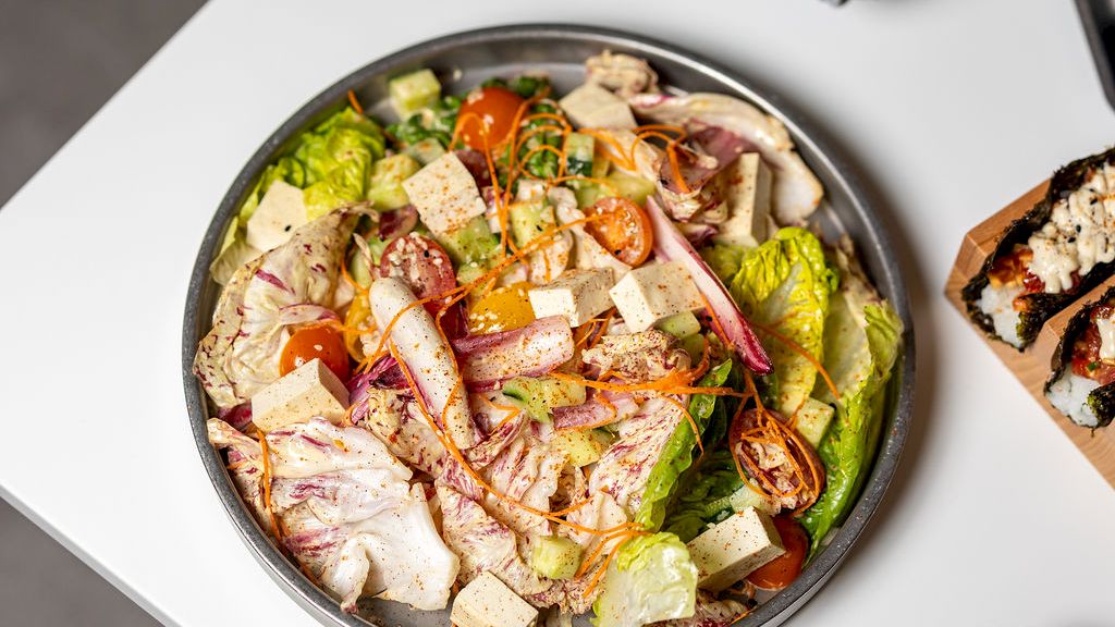 A fresh salad bowl with mixed greens, tomatoes, cucumbers, cheese, and chunks of chicken on a white table with a grilled slice of bread nearby.