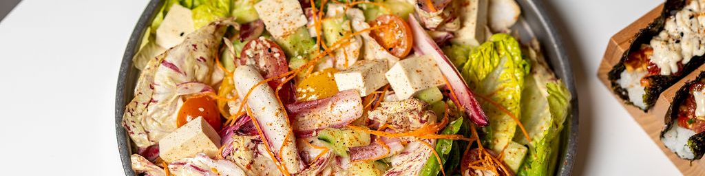 A fresh salad bowl with mixed greens, tomatoes, cucumbers, cheese, and chunks of chicken on a white table with a grilled slice of bread nearby.