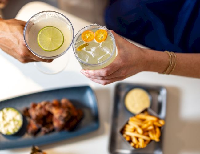 Two people clink glasses of citrus cocktails with lime and orange slices, beside grilled food, fries, and sandwiches on a picnic table.
