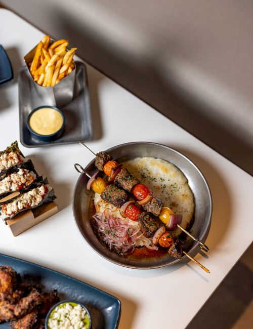 A skillet of grilled meat with veggies on skewers, plus fries with dip, and breaded bites on plates nearby, all on a light table.
