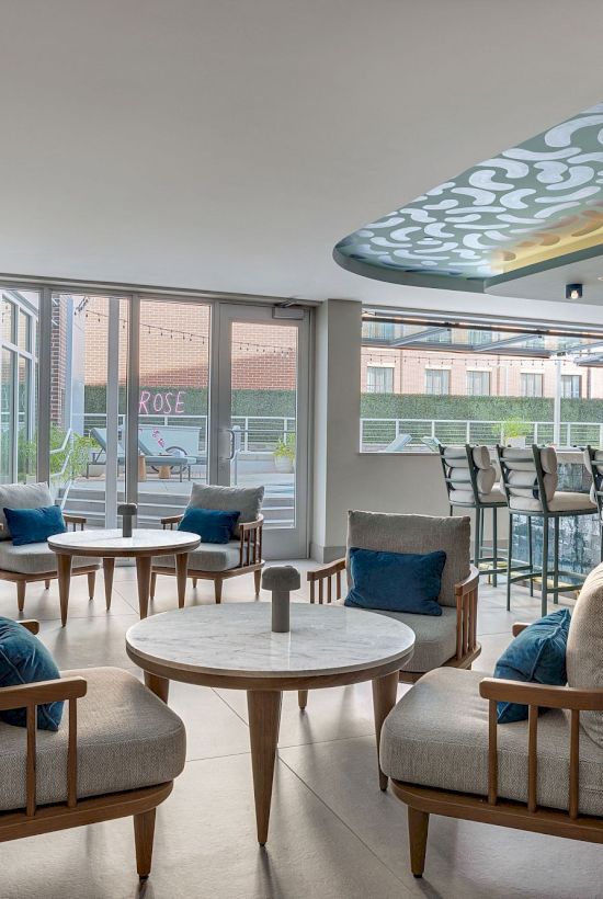 Modern lounge with cushioned chairs and round tables, bar stools by a water feature, and a patterned ceiling over a patio view.