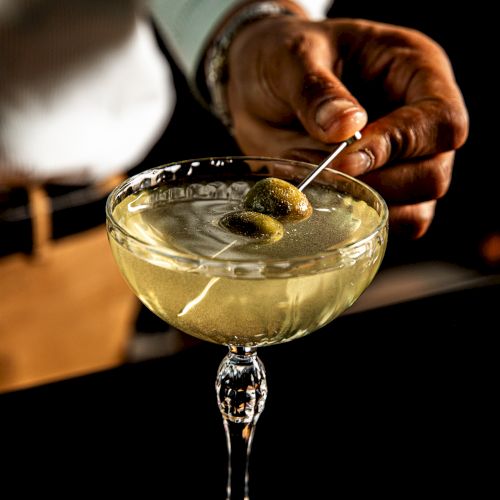 A bartender pours a pale cocktail into a coupe glass, garnished, with a lime wedge; a drink appears to be a whiskey sour or similar.