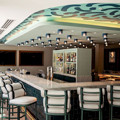 A modern hotel bar lounge with a marble counter, stools, a lit back bar, and a curved ceiling with spotlit lamps.