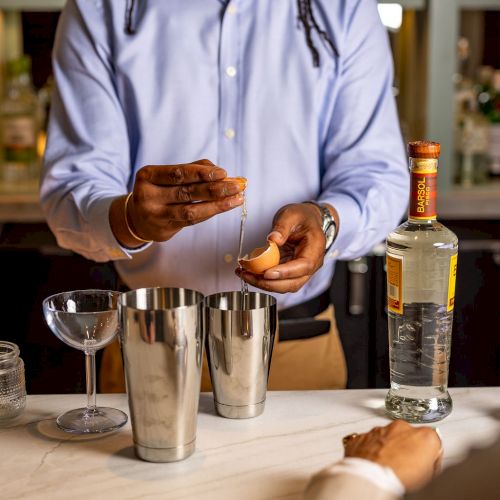 A bartender pours ice into a shaker behind a bar, with bottles and tools on the counter.