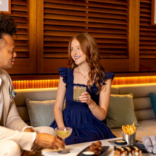 Two people enjoying drinks and snacks on a lounge couch, chatting warmly in a stylish setting with plates, fries, and candles, inside a chic lounge.