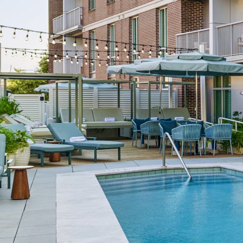 A modern brick apartment patio with a blue pool, teal lounge chairs, umbrellas, string lights, and garden plants, for relaxing outdoors.