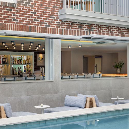 A modern brick-and-concrete patio with an outdoor bar window, string lights, and blue cushioned loungers beside a swimming pool.