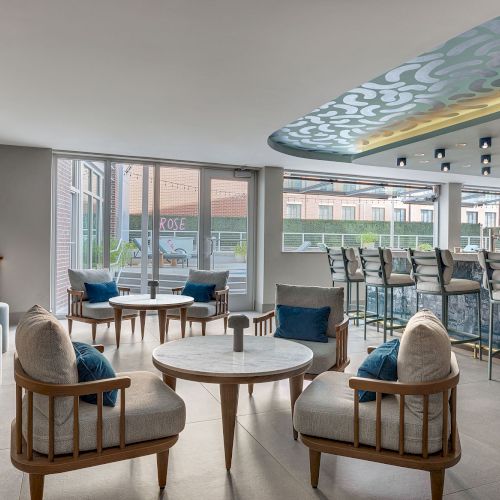 A modern indoor lounge with comfy chairs and a round table, glass walls, and a patterned ceiling canopy with built-in lights.