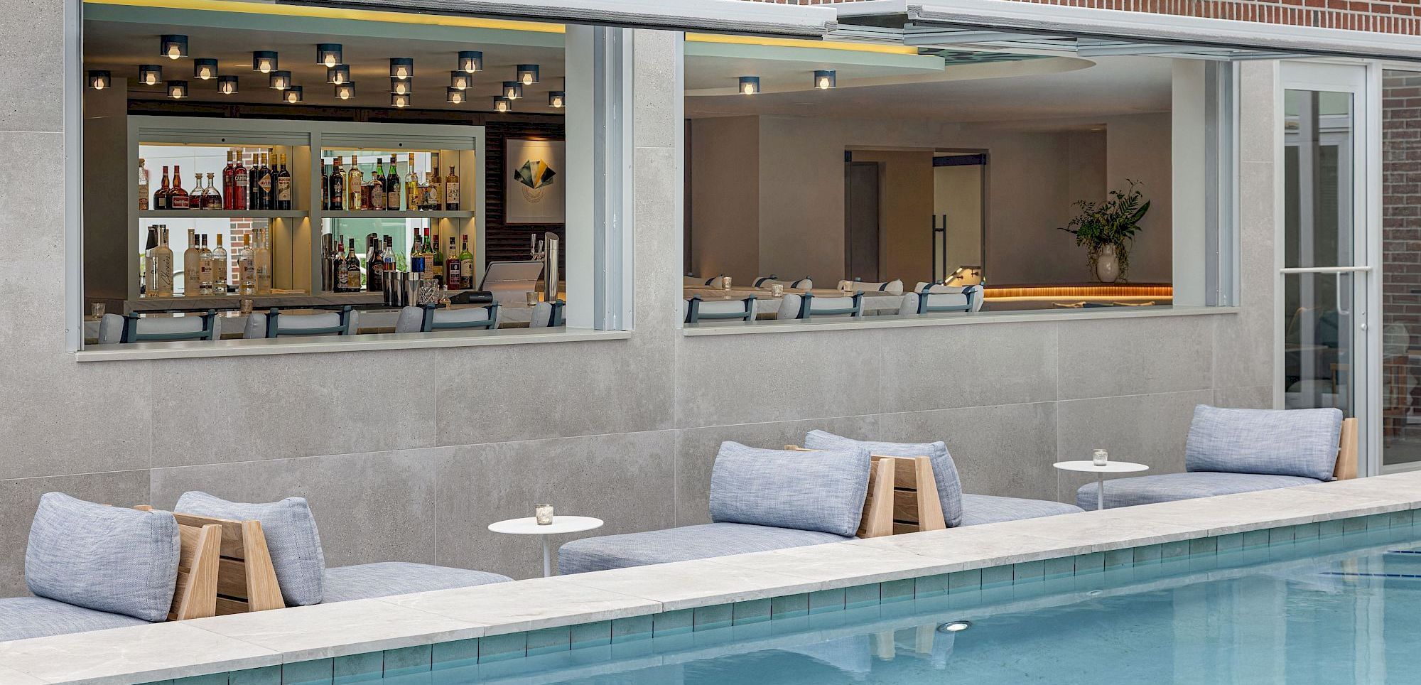 A modern brick building poolside bar with an open service window, bottles on shelves, lounge seating, and a reflecting swimming pool.