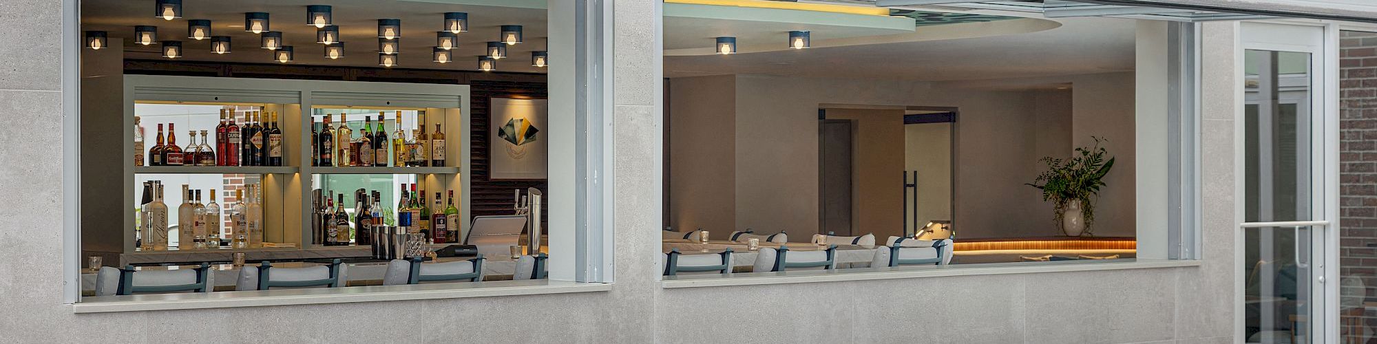 A poolside patio with cushioned lounge seating and an outdoor bar window stocked with bottles, set against a brick building.